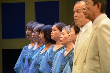 白色のスーツを着た男性と水色のドレスを着た女性が横一列に並んで歌を歌っているシーンの写真