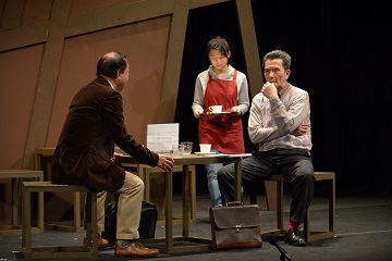 2名の男性が話し込んでいるテーブルに赤色のエプロンを着用した女性がコーヒーを運んできているシーンの写真