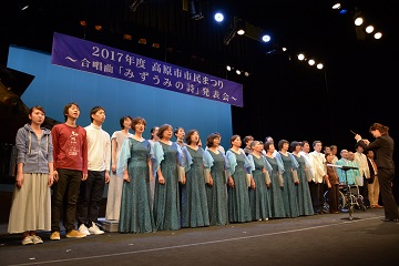 水色のドレスを着た来た女性たちと私服姿の若い人たちがステージに立って歌を歌っているシーンの写真