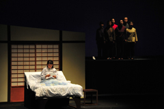 右側の高い位置に立っている男女と左側のベッドの上で手紙を読んでいる女性のシーンの写真