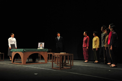 女性の遺影を乗せたテーブルを挟んで間隔をあけて立っている男女、右側の6名の男女が並んで立っているシーンの写真