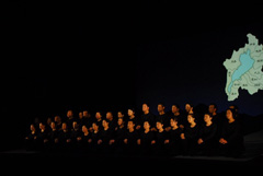 右側に滋賀県の地図が浮かび上がっている照明を落としたステージ上で全身黒色の服を着た2列に並んでいる人達が立っている写真
