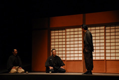 左側に間隔をあけて座っている男性と右側に立っている男性が話をしているシーンの写真