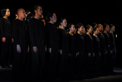 ステージ上に全身黒色の服を着た人達が2列に並んで立っているのを左斜めから撮影した写真