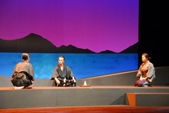 空がピンク色に染まっているバックの前で着物を着て正座した女性と男性が向かい合って話しているのを右側に間隔をあけて座って見ている女性の劇のシーンの写真