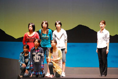 前列は座り後列は立ち2列に並んでいる女の子と右側に立っている1名の女性が島と海をバックに横一列に並んで立っている劇のシーンの写真