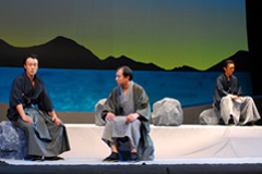 奥の石に1名の男性が座り、左手前の石に2名の男性が並んで座って話をしているシーンの写真