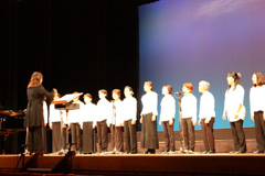 白と黒色の衣装を着た人たちが横一列に並んで指揮に合わせて歌を歌っている写真