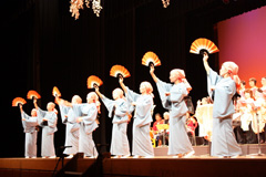 後方の演奏と歌に合わせて横一列に並んだ着物姿の女性たちが扇子を右手で持って踊っている様子の写真