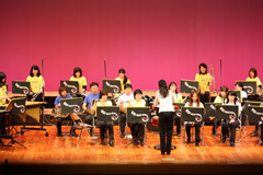 指揮に合わせて黄色と紫色のシャツを着た人たちが演奏を行っている様子の写真