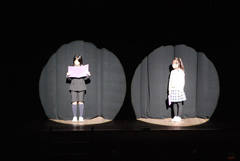 ステージ上に間隔をあけて照明に照らされた2名の女の子が立ち、左の女の子が両手に持った本の様な物を見ながら言葉を発している様子の写真