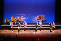 後方で黄色の法被を着た人の太鼓や三味線の演奏に合わせてピンクと黒色の衣装を着た6名の女性が踊っている様子の写真