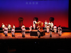 ステージの中央に設置された小さな舞台の上で叩いている太鼓の演奏に合わせて着物を着て踊っている女性たちの写真