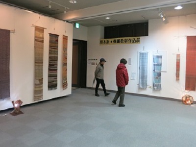 様々な色や模様があしらわれた作品が壁に展示されているのを2名の男性と女性が見に来ている写真