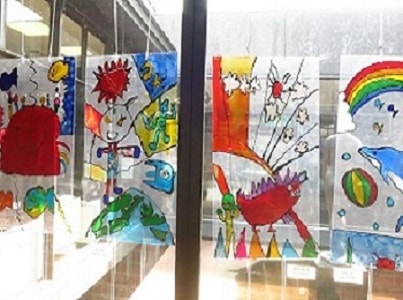 窓ガラスに子供たちが透明の板に描いたカニや虹の下で飛び跳ねているイルカなどの作品が貼られている写真