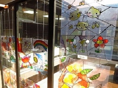 窓ガラスに子供たちが透明の板に描いた猫の顔や花などの作品が貼られている写真