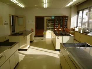 前方に食器棚、右側に窓があり、洗い場とガスコンロが付いた作業台が設置してある調理実習室の写真