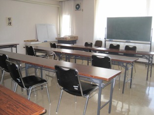 黒板があり、左側にホワイトボードが立てかけられ、長机と黒い椅子が置かれている会議室の写真