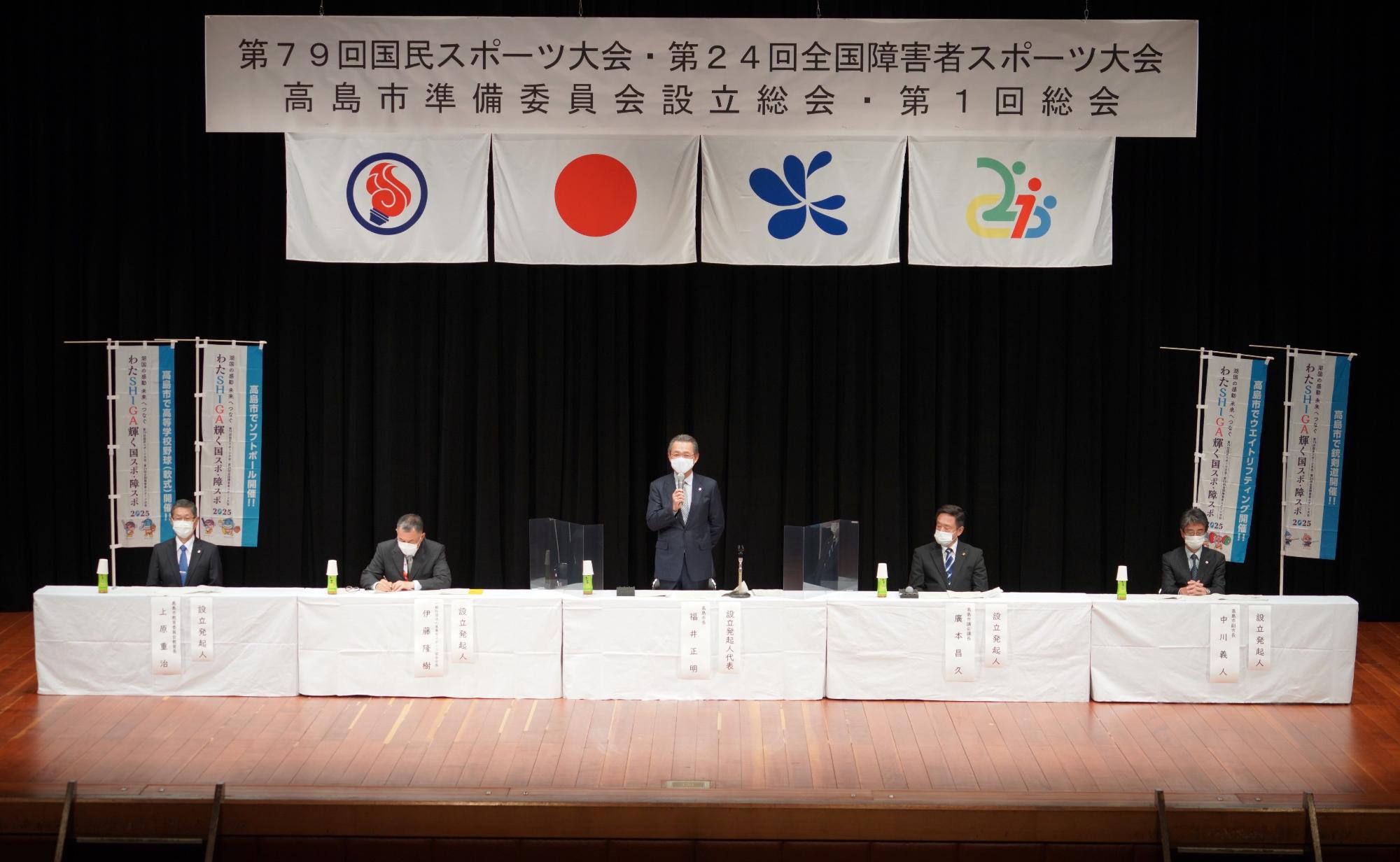 壇上に設置された机と椅子に準備委員会の方々が座り、中央の福井市長が立ちマイクを使って話をしている様子の写真