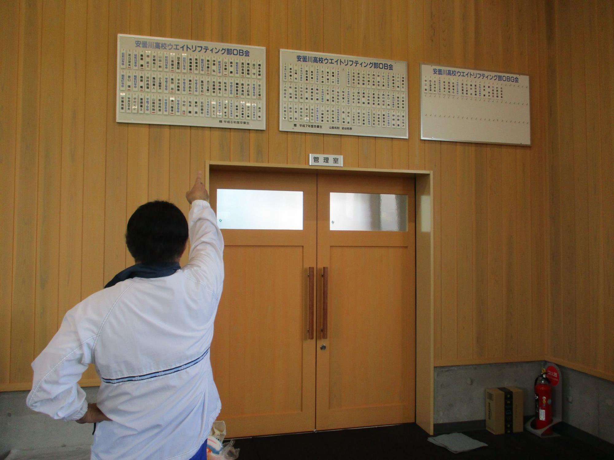 管理室入り口の壁上に掲げた安曇川高校ウエイトリフティング部OBOGの名前札を西村さんが右手の人差し指で差している後ろ姿の写真