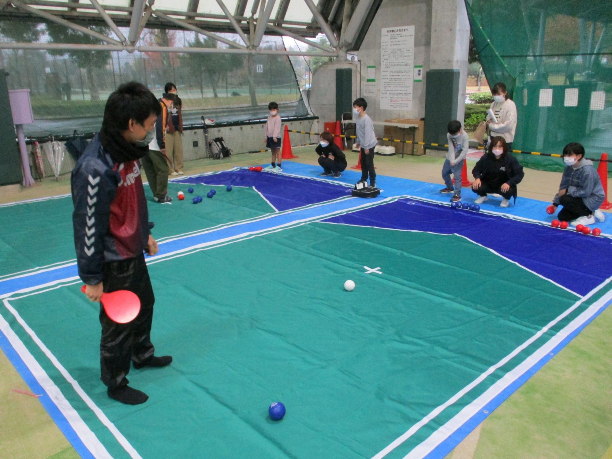 中央の印の傍に白色のボールがあり、左側の卓球のラケットの様な道具を右手で持って立っている男性と右側に赤色のボールを持って座っている女性が向かい合っている写真