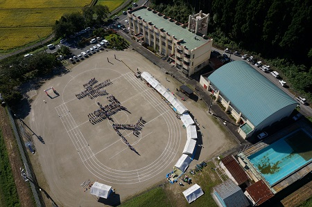校庭に人文字でマ北小と書かれているマキノ北小を上空から撮影した写真