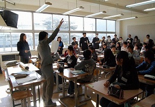 壁側に関係者が立ち、先生が右手を上げて生徒に問いかけている小学6年生授業の様子の写真