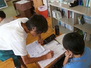 先生が机に置かれた宿題をチェックするのを見ている生徒の夏休み学習会の様子の写真
